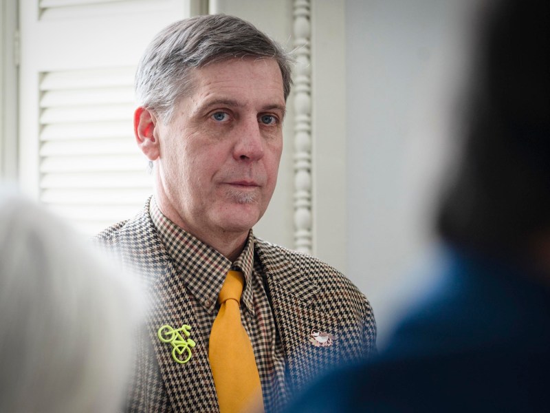 A man in a brown checkered jacket and yellow tie sits indoors, wearing a bicycle-shaped pin on his lapel.