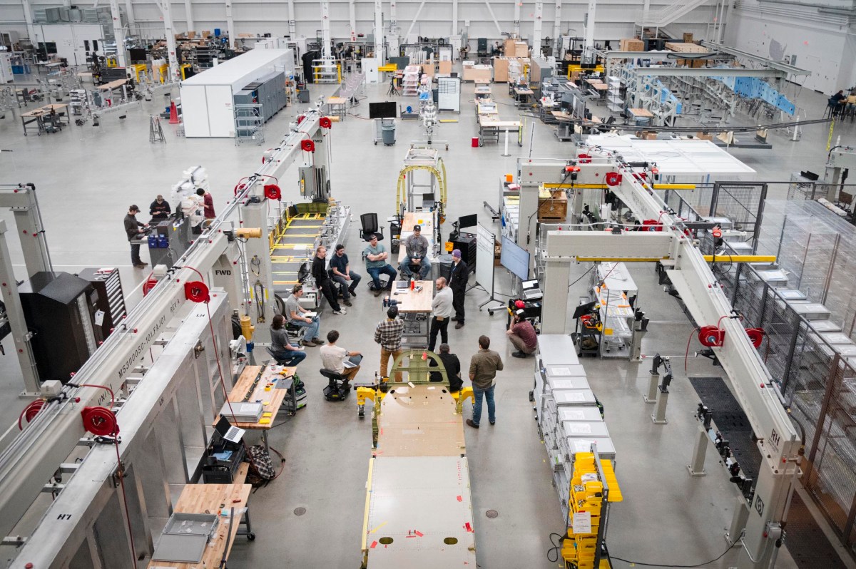 Overhead view of a spacious factory floor with workers gathered around a central assembly line, surrounded by machinery, workstations, and industrial equipment.