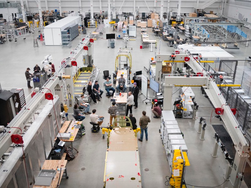 Overhead view of a spacious factory floor with workers gathered around a central assembly line, surrounded by machinery, workstations, and industrial equipment.