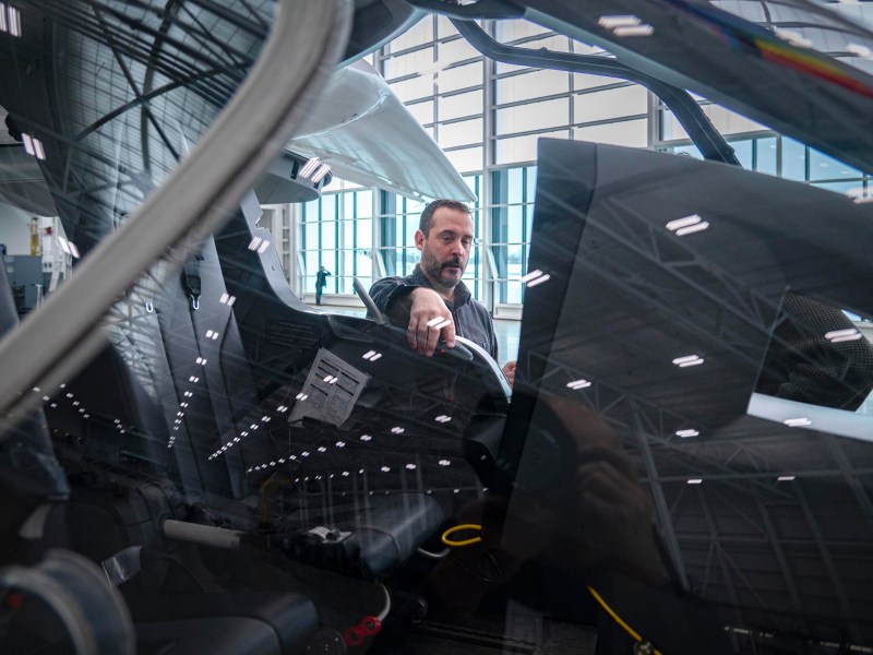 A man stands next to a modern aircraft cockpit inside a hangar, with reflections of the hangar lights visible on the glass.