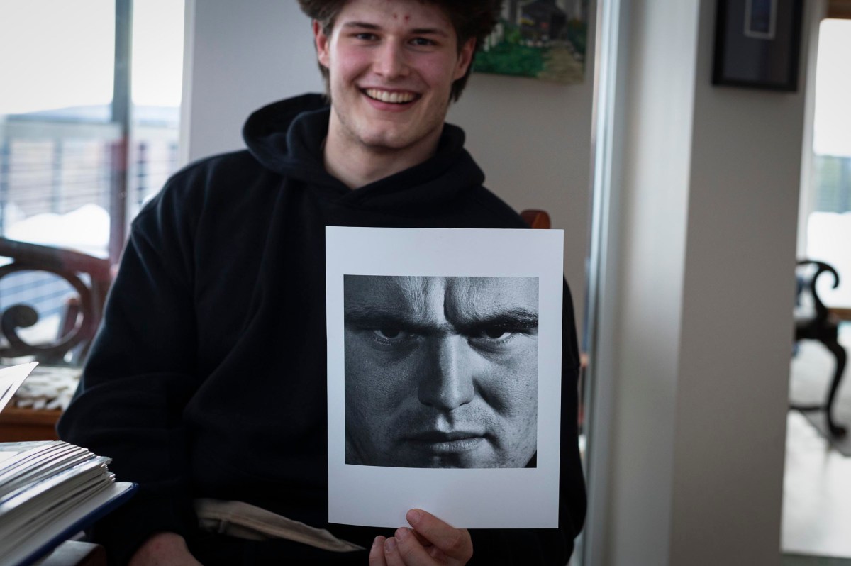A smiling person in a black hoodie holds a paper with a large black-and-white photo of a serious man's face.