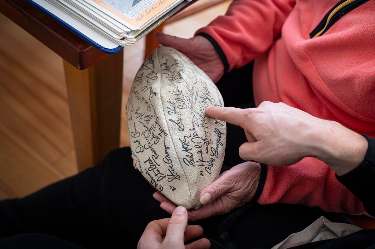 Two people hold and point at a football covered in handwritten signatures, with a book or album visible on the table nearby.
