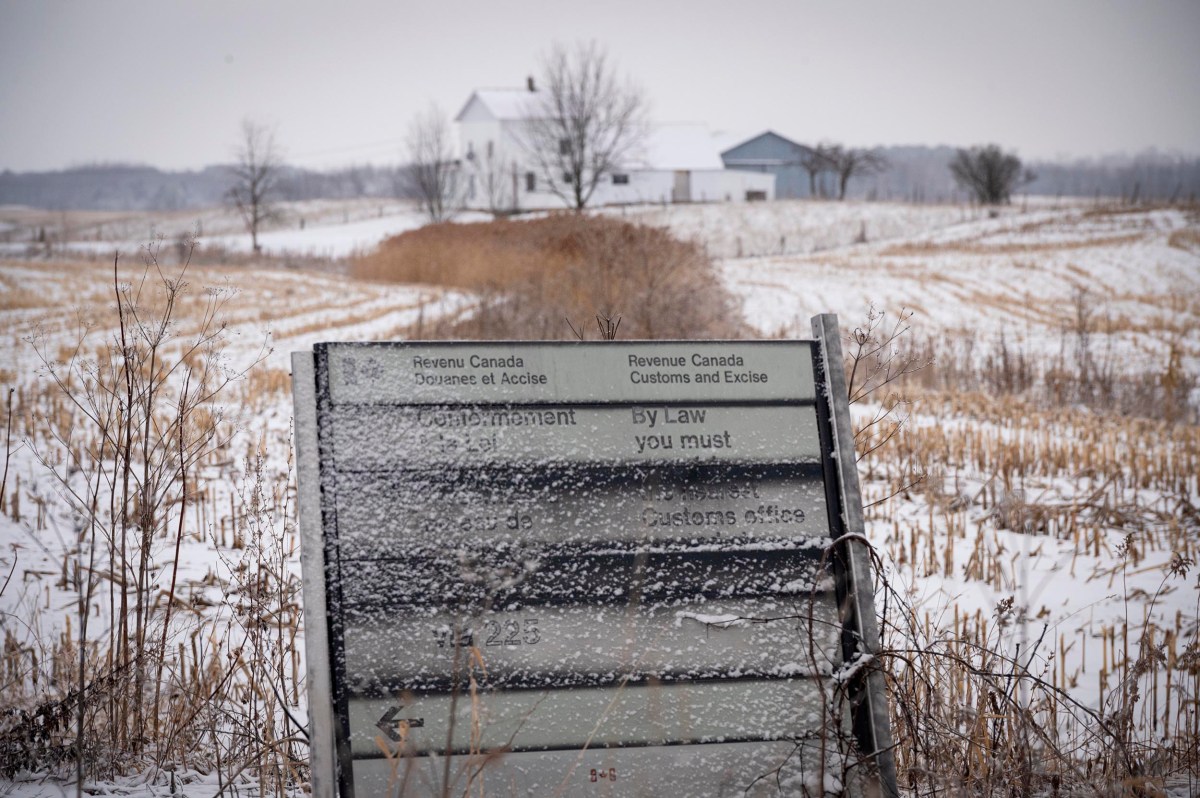 A weathered, ice-covered Customs and Excise sign stands in a snowy, rural field with a white farmhouse and barns in the background.