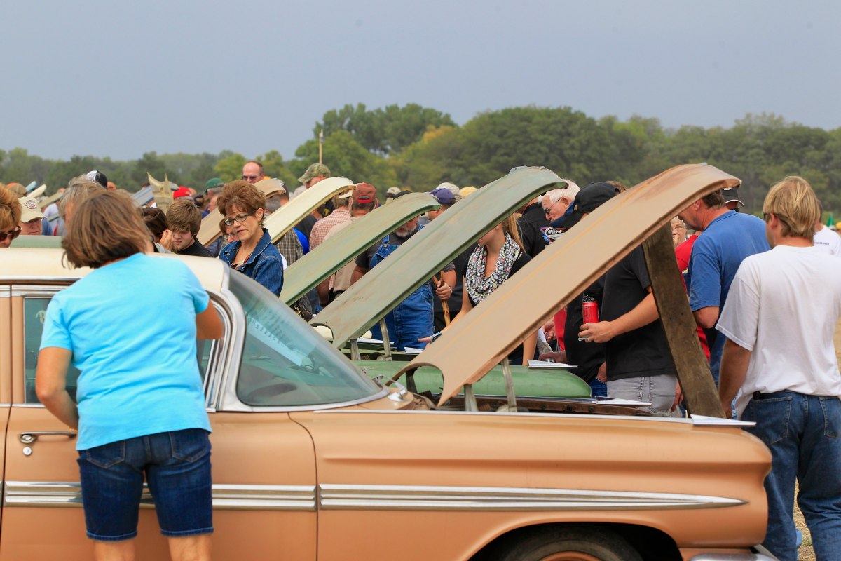 People gather outdoors around several classic cars with open hoods, inspecting engines and parts. Trees and a cloudy sky are visible in the background.