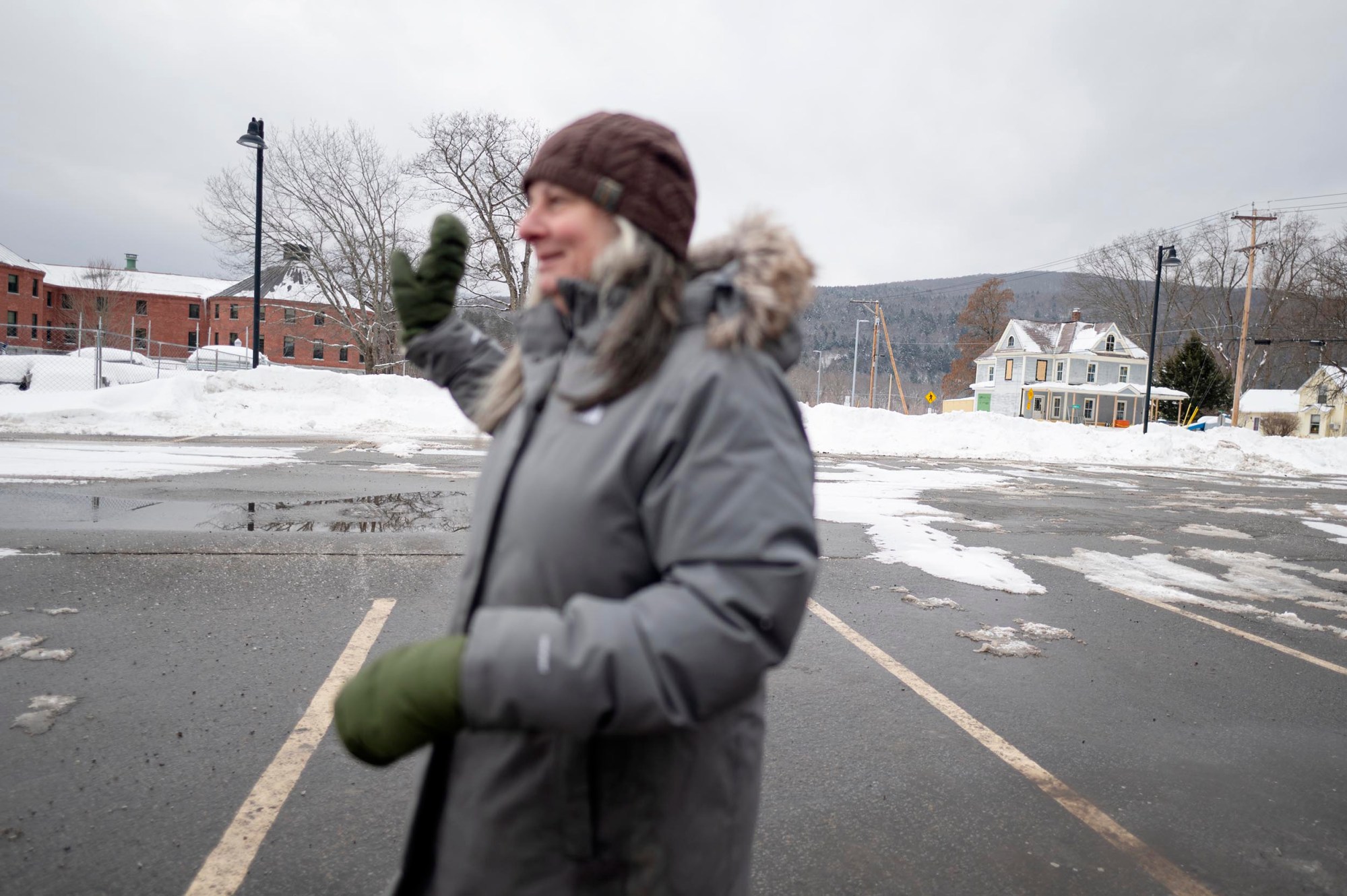 A person in a gray winter coat, green mittens, and brown hat stands in a snowy, empty parking lot, with buildings and mountains in the background.