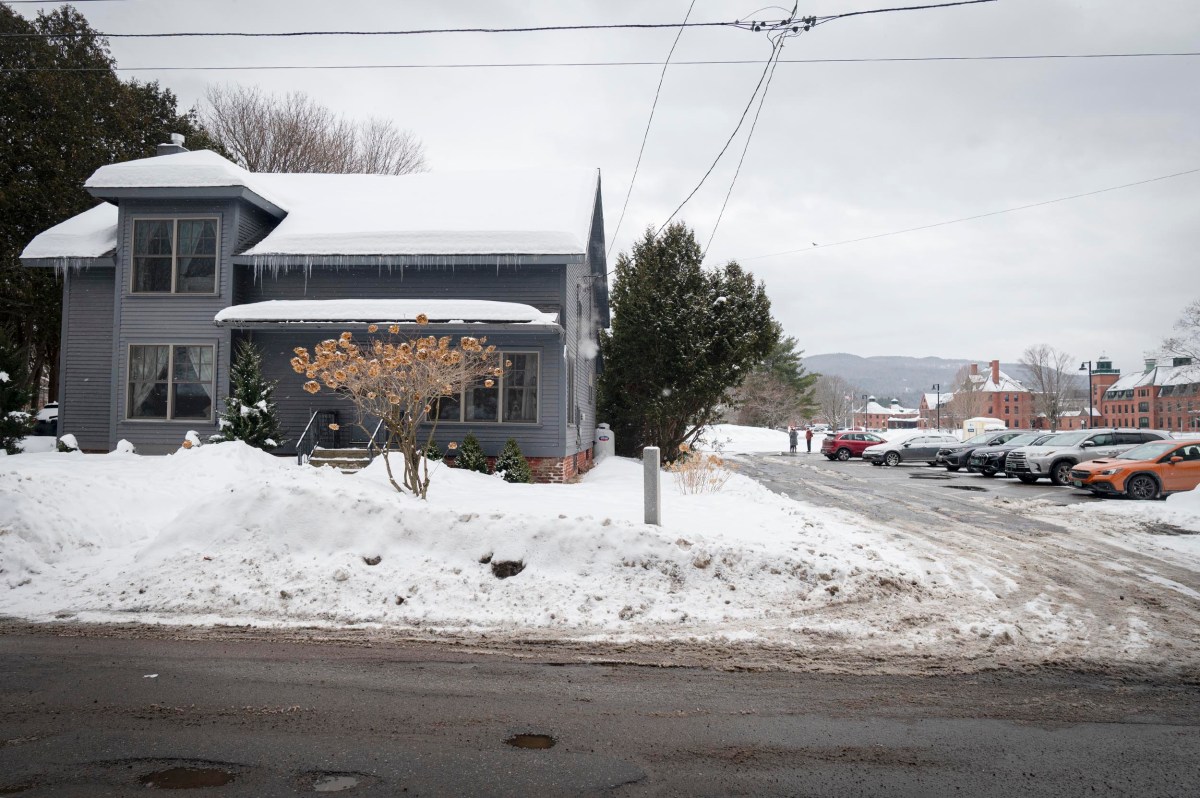 A gray house with icicles on the roof stands beside a snow-covered yard and driveway lined with parked cars on a cloudy winter day.