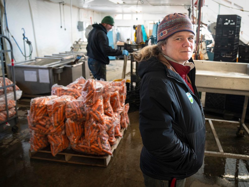A woman in a winter jacket stands in a produce processing facility with bags of carrots stacked on a pallet; a man works in the background.