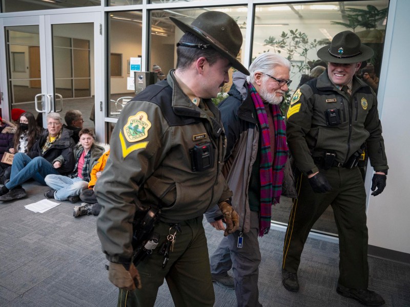 Two police officers escort an older man through a hallway as several seated protesters watch from the floor against a glass wall in the background.