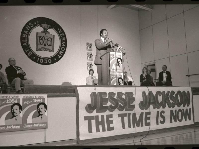 Jesse Jackson speaks at a podium on a stage with large campaign signs reading "JESSE JACKSON THE TIME IS NOW" and "REGISTER & VOTE 1984." Audience and campaign posters visible.