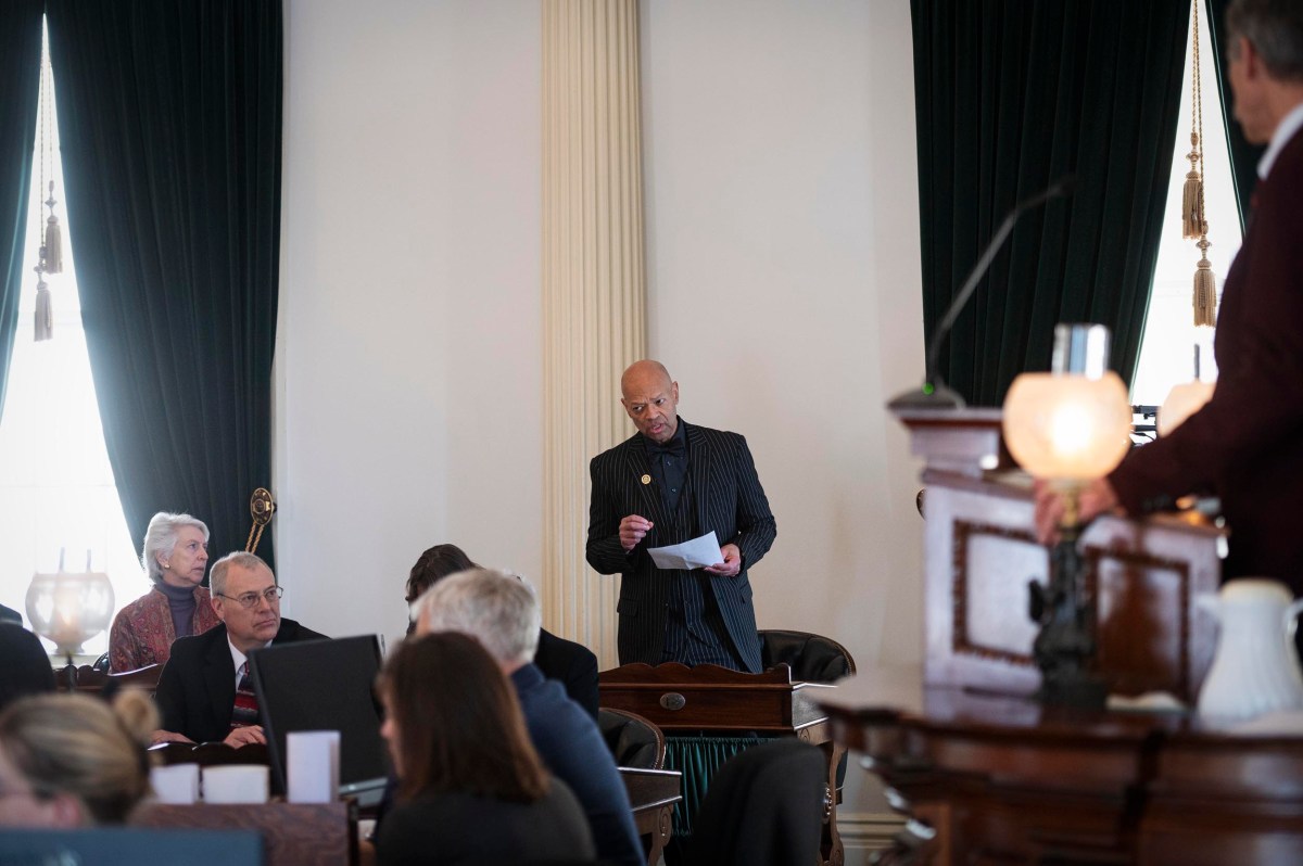 A man stands holding papers while addressing a seated group in a formal room with dark curtains and large windows.