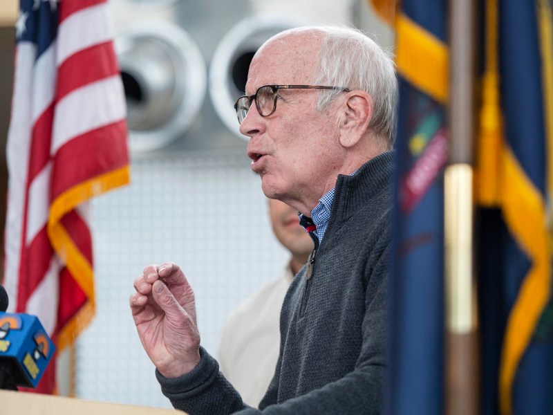 An older man with white hair and glasses speaks at a podium, with American and other flags in the background. Microphones are positioned in front of him.