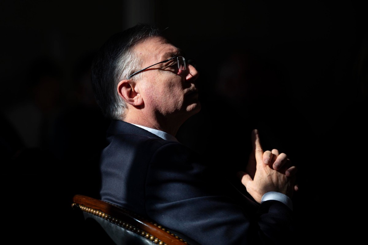 A man in a suit and glasses sits in profile with his hands clasped, illuminated by a beam of light against a dark background.