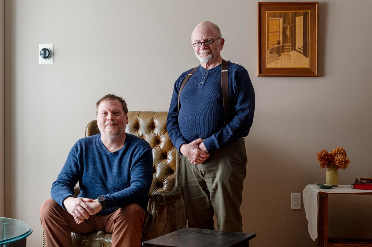 Two men indoors; one is seated in an armchair while the other stands beside him. A painting, a table with dried flowers, and a thermostat are visible in the background.