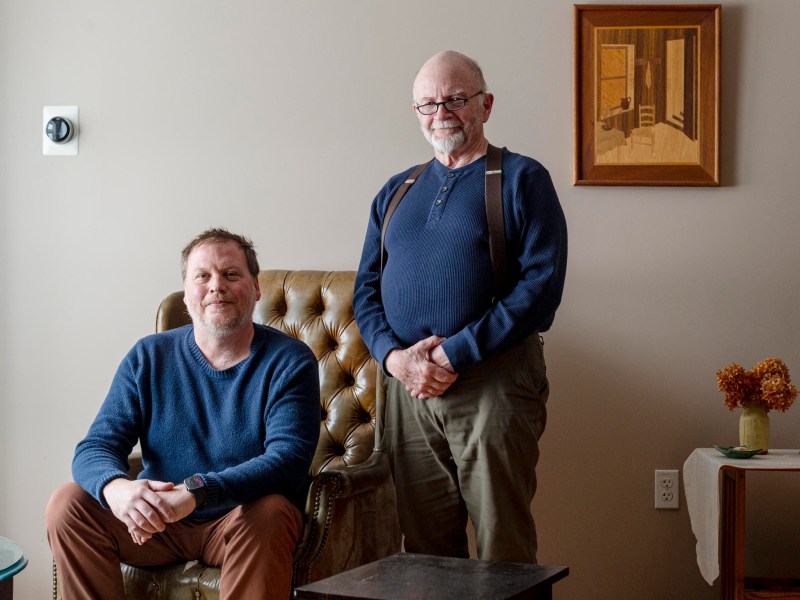 Two men indoors; one is seated in an armchair while the other stands beside him. A painting, a table with dried flowers, and a thermostat are visible in the background.