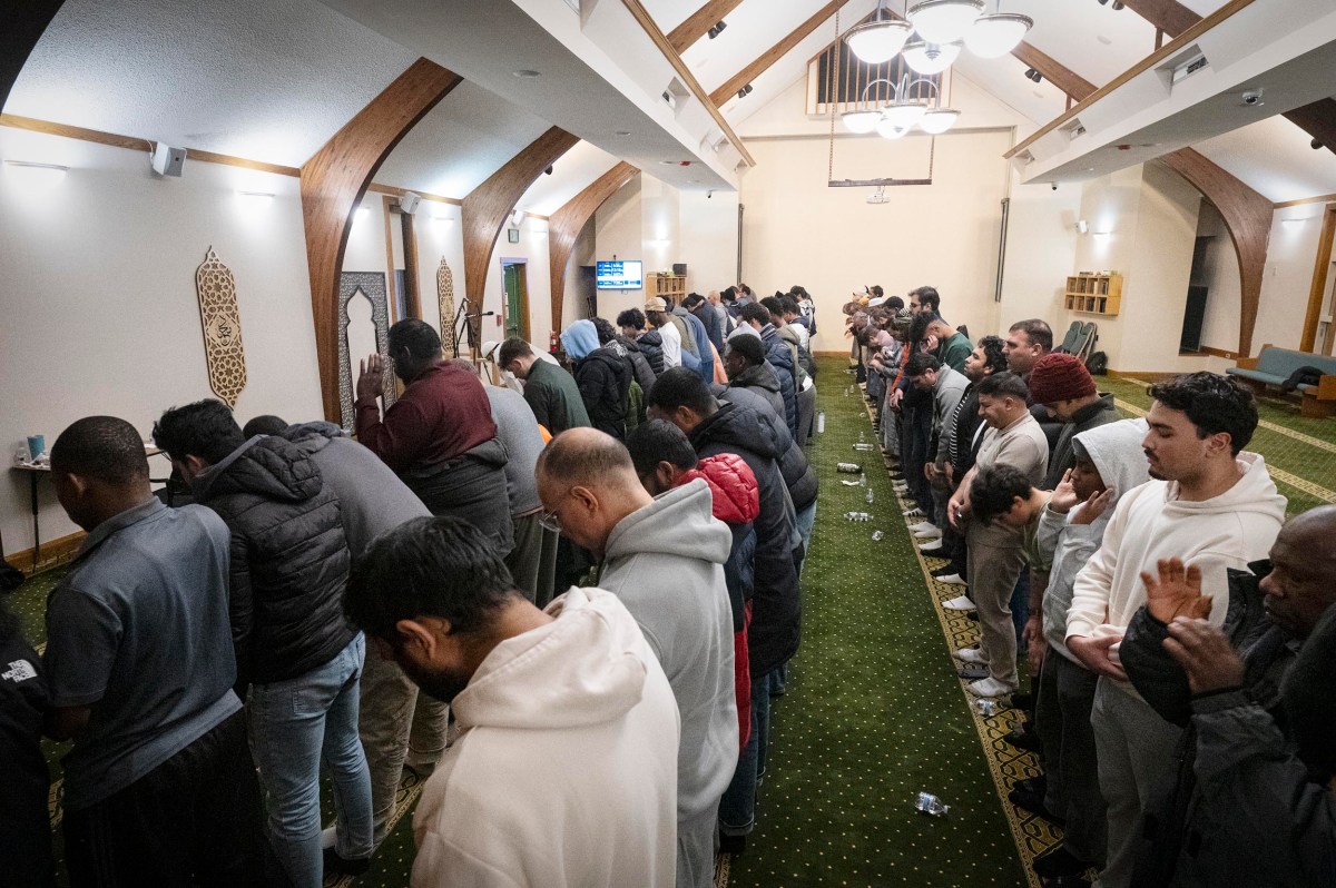 A group of people stand in rows, facing forward with hands raised in prayer, inside a mosque with patterned green carpet and arched wooden beams.
