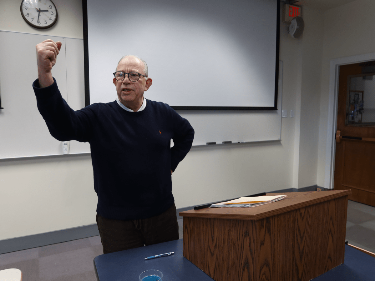 An older man wearing glasses stands at a lectern in a classroom, raising his right fist and speaking, with a projection screen and whiteboards behind him.