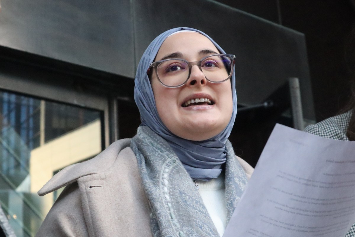 A person wearing glasses and a light blue hijab speaks while holding papers, standing in front of a building.