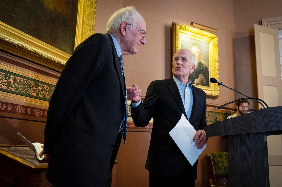 Two men in suits stand together in a room with framed portraits; one speaks at a podium while the other listens and holds papers.