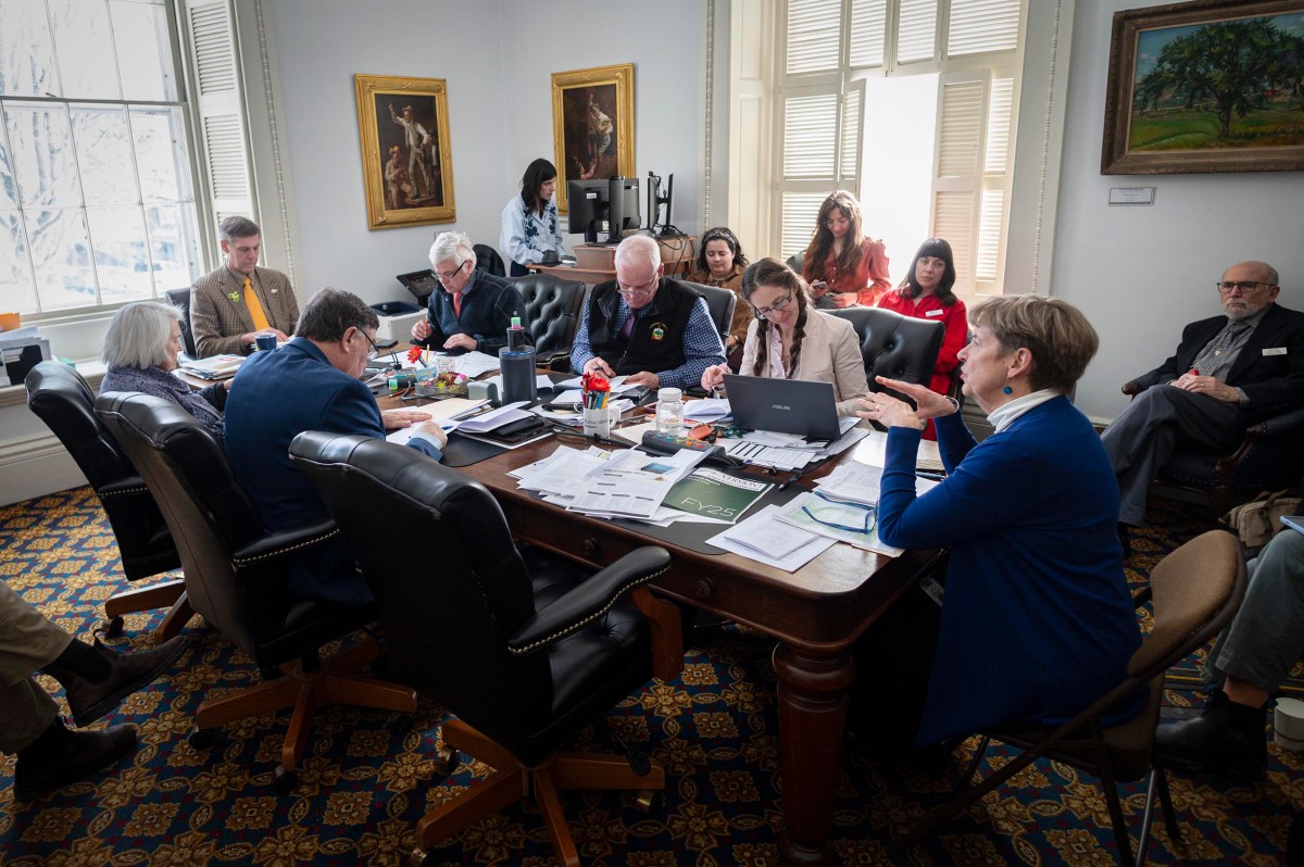 A group of people sits around a large table covered with papers and laptops in a meeting room, while several others observe from chairs along the wall.