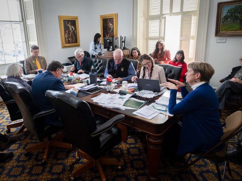 A group of people sits around a large table covered with papers and laptops in a meeting room, while several others observe from chairs along the wall.
