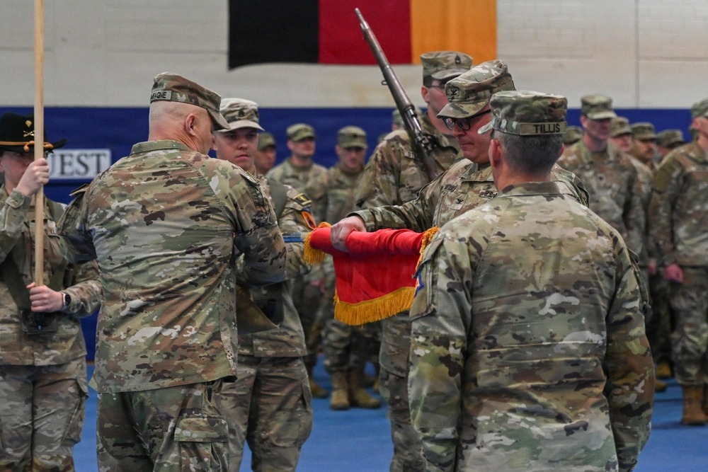A group of soldiers in uniform participate in a flag ceremony indoors, with one soldier handing a flag to another while others stand in formation.