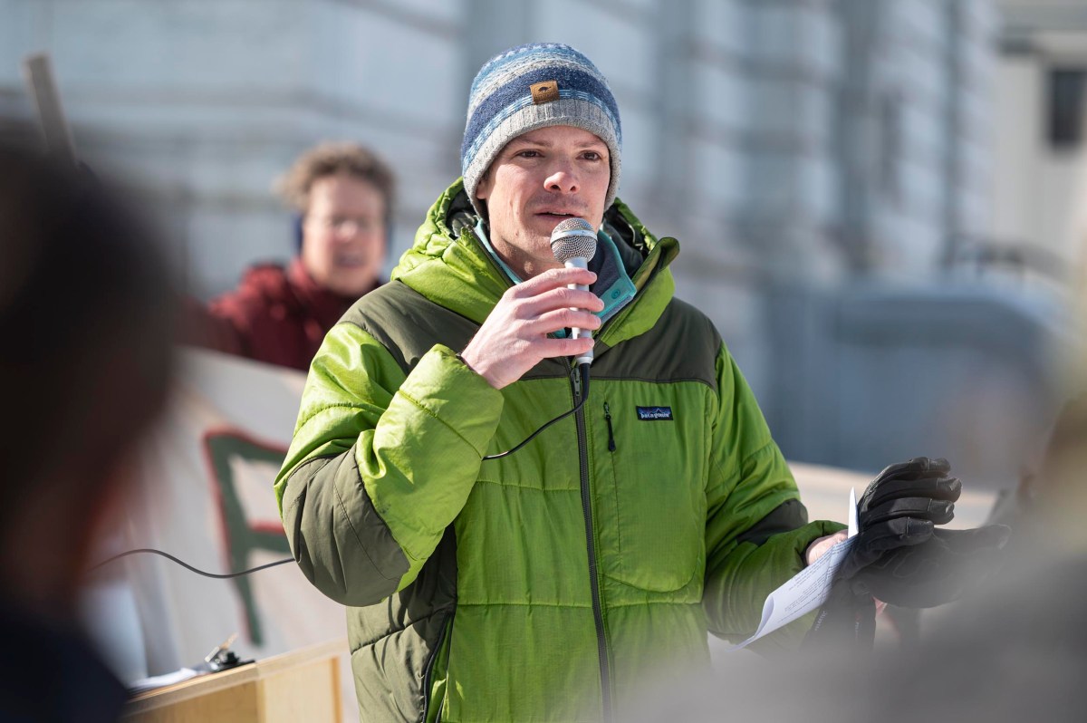 A person in a green jacket and blue beanie speaks into a microphone outdoors, holding papers, with another person and a banner visible in the background.