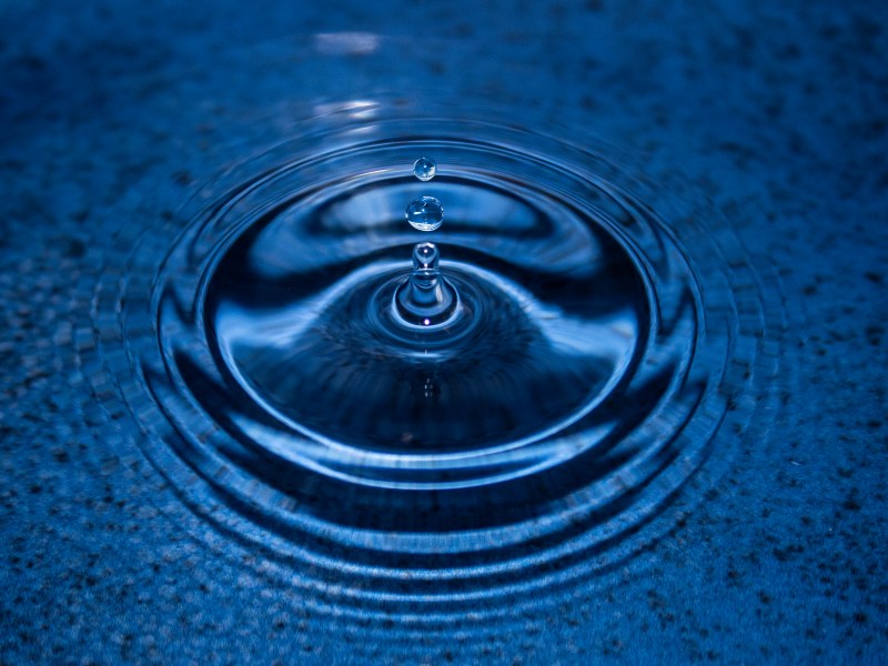 Close-up of water droplets falling into a blue surface, creating circular ripples and splashes around the point of impact.