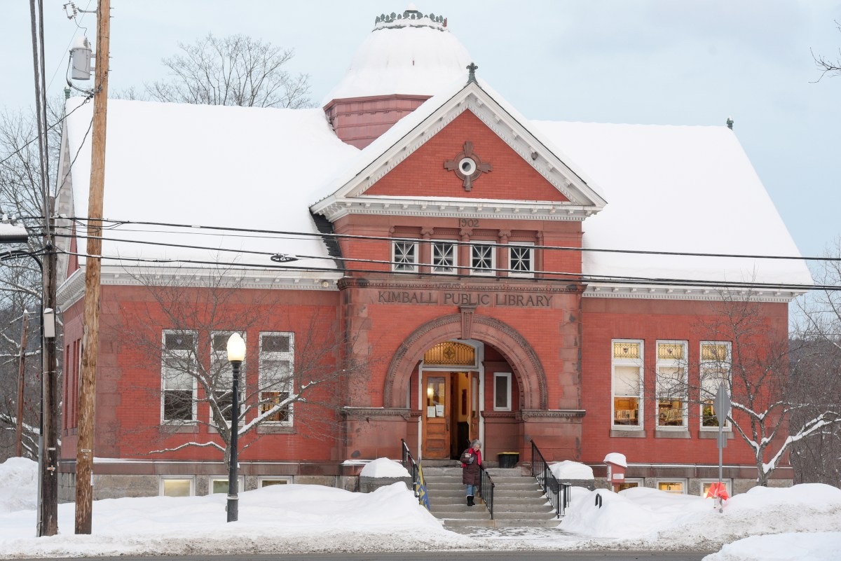 Red brick public library building with arched entrance and snow-covered roof, surrounded by snow-covered ground and leafless trees in winter.