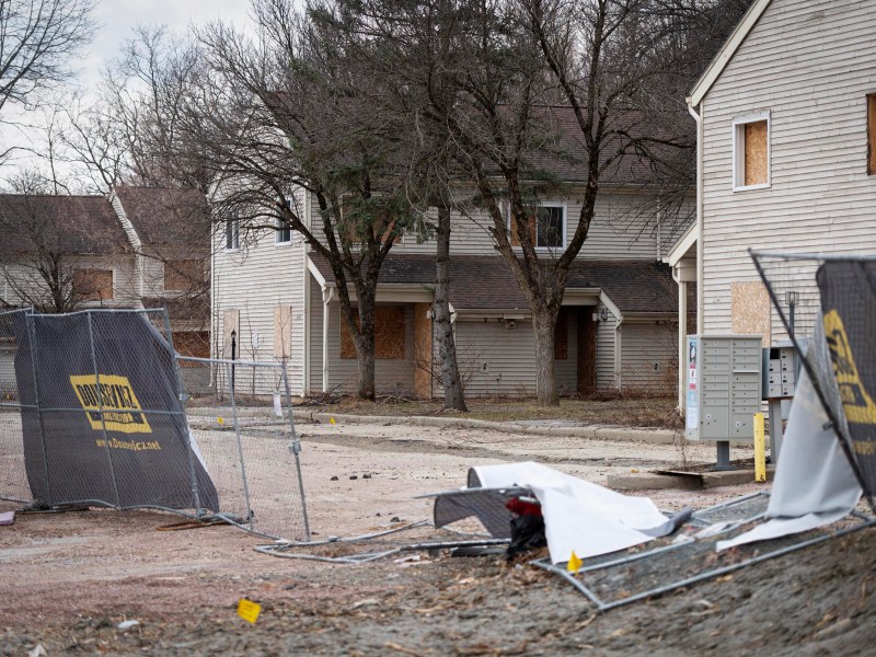 Two houses with boarded-up windows sit behind a damaged construction fence and debris on a cloudy day.
