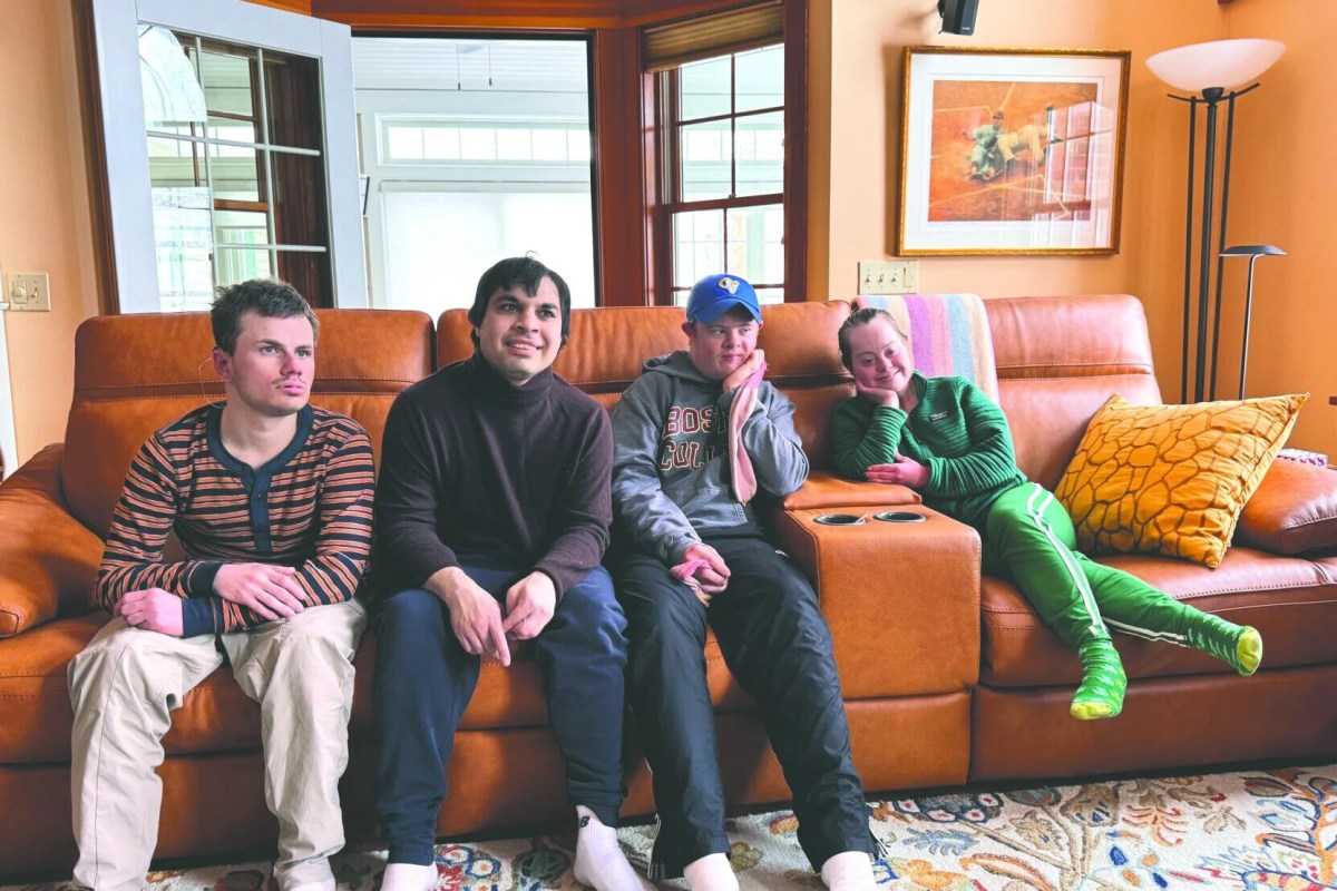 Four people sit together on a brown leather couch in a brightly lit living room with orange walls and a framed painting above them.