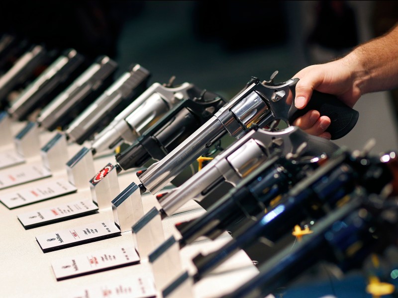 Several handguns are displayed in a row on a counter, with a person holding one revolver; information cards are placed in front of each firearm.