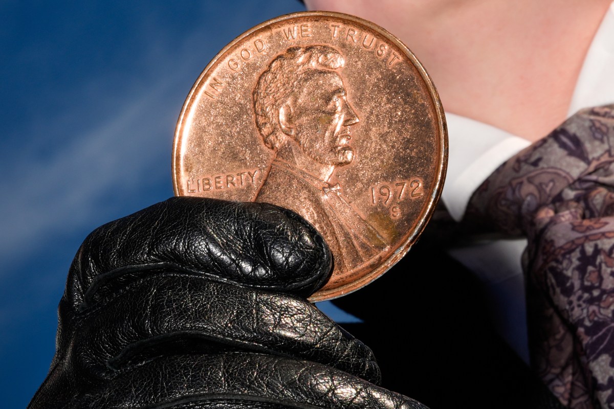 A person wearing a black leather glove holds an oversized 1972 US penny with Abraham Lincoln's profile against a blue background.