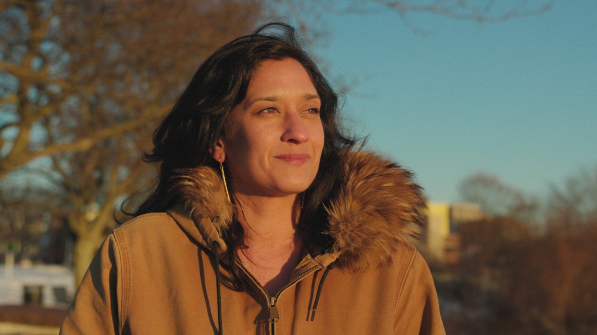 A woman in a tan coat with a fur-lined hood stands outdoors, looking into the distance, with trees and buildings blurred in the background.