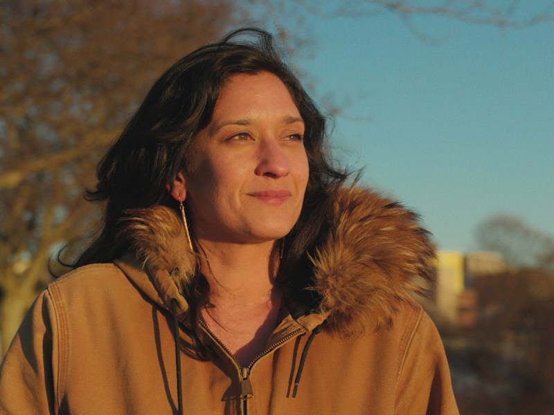 A woman in a tan coat with a fur-lined hood stands outdoors, looking into the distance, with trees and buildings blurred in the background.