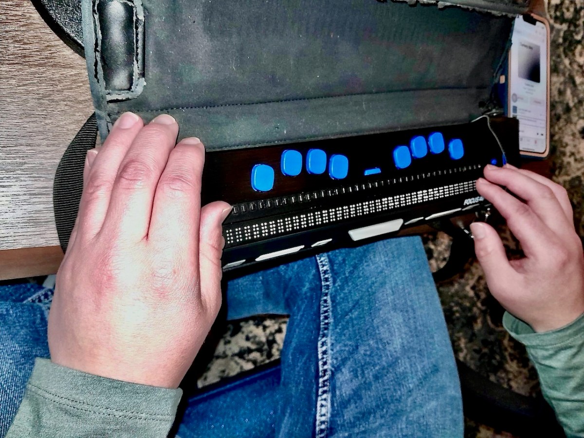 A person uses a braille display device with blue keys, reading with both hands while a smartphone rests nearby.