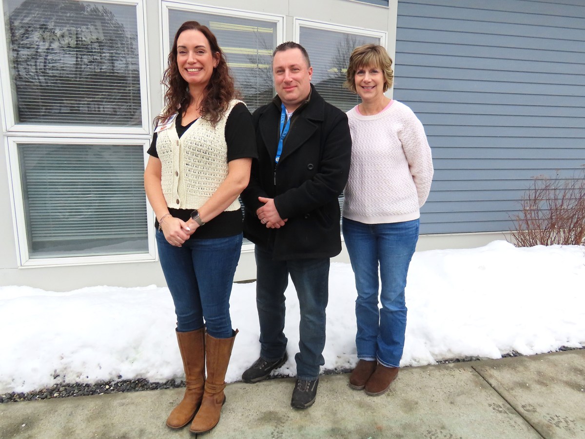 Three adults stand outside on a snowy sidewalk in front of a building with large windows and blue siding.