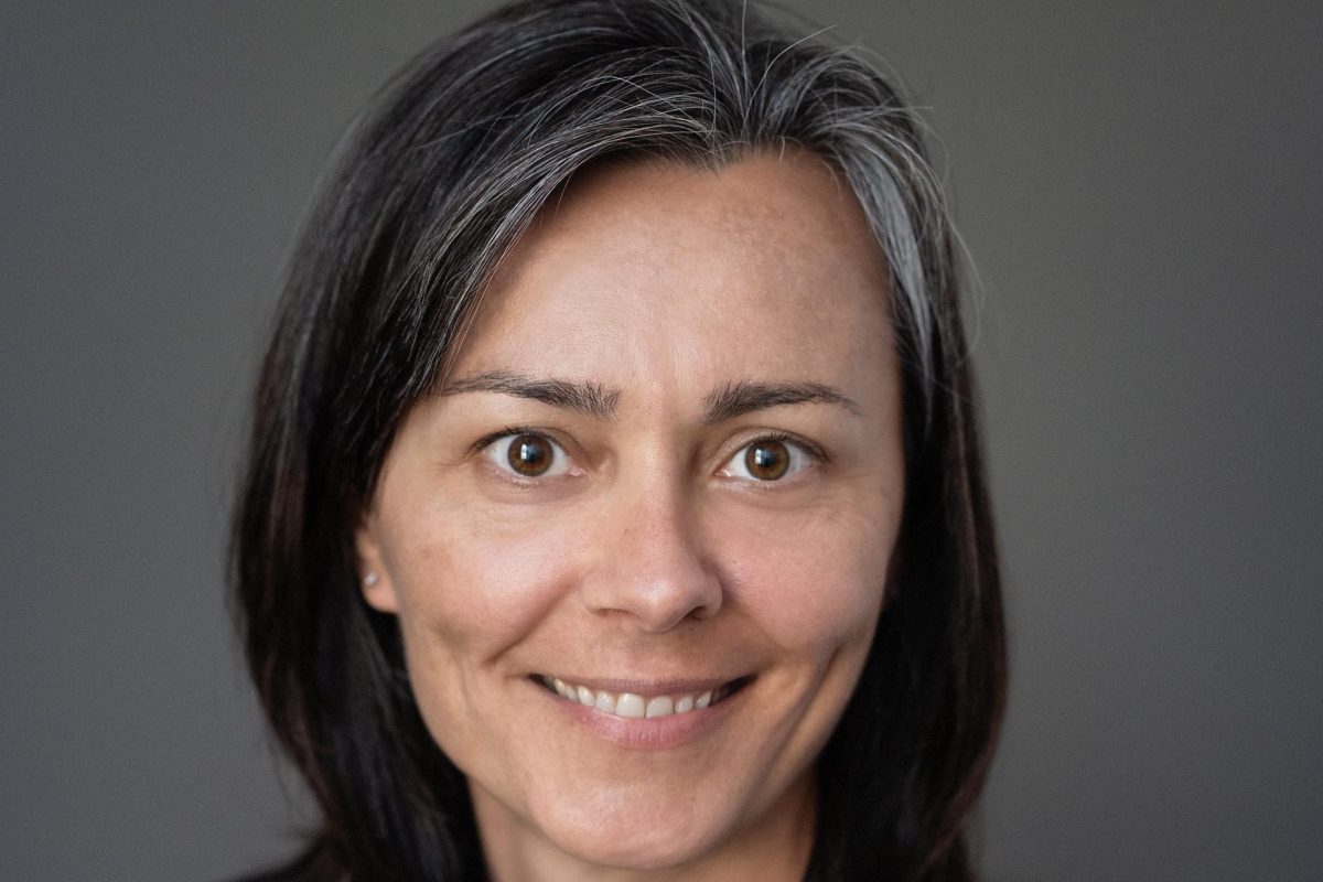 A woman with straight dark hair and streaks of gray, wearing a black blazer and patterned top, smiles at the camera against a plain gray background.