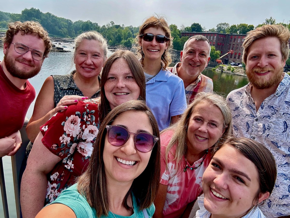 A group of nine adults pose for a selfie outdoors in front of a river, trees, and a red brick building on a sunny day.