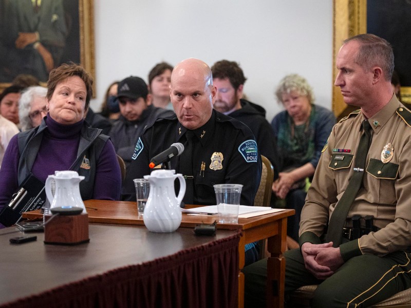 Three people, including two uniformed officers and a woman, sit at a table with microphones and water pitchers during a formal meeting or hearing, with an audience seated behind them.
