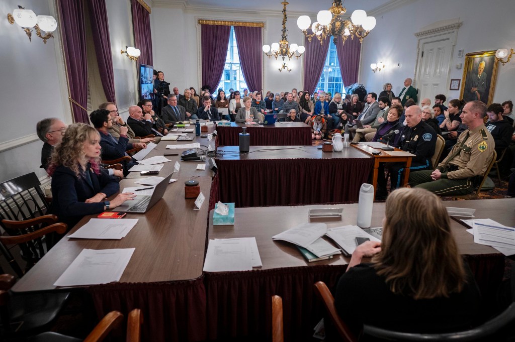 A government meeting in a formal room with officials seated at tables and an audience observing; documents and microphones are on the tables.