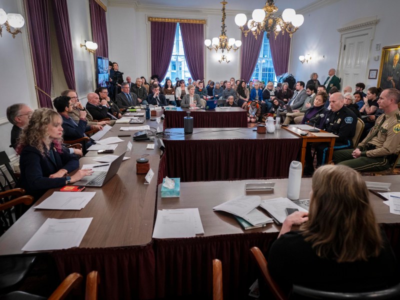 A government meeting in a formal room with officials seated at tables and an audience observing; documents and microphones are on the tables.