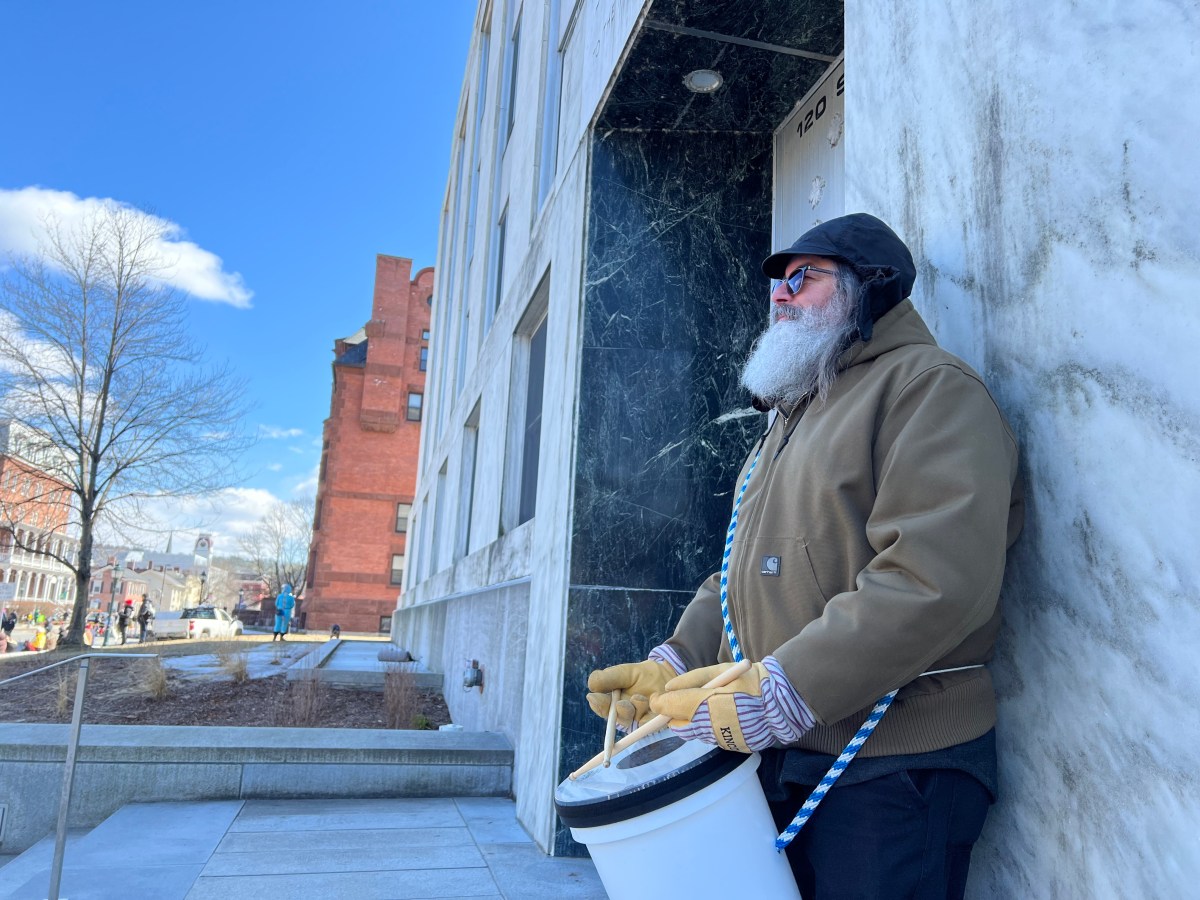 A man with a white beard, wearing a brown jacket and gloves, stands outside a marble building holding a white bucket.