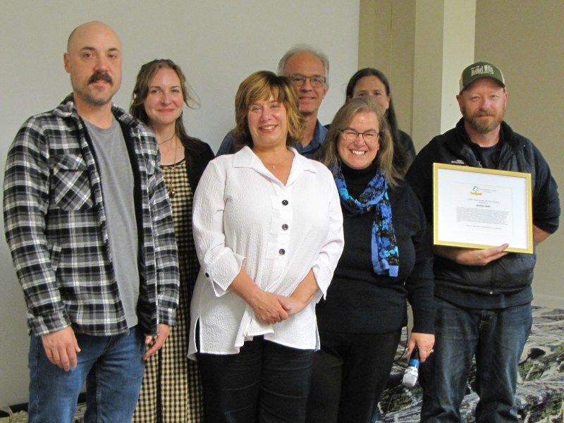 Seven adults pose indoors; one holds a framed certificate. They are standing in front of a blank wall, smiling at the camera.