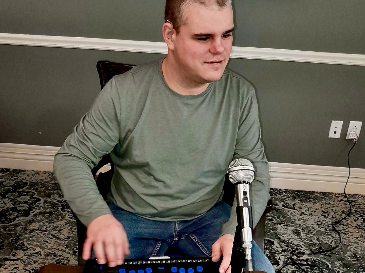 A man in a green long-sleeve shirt sits at a table using an electronic braille device, with a microphone positioned in front of him.