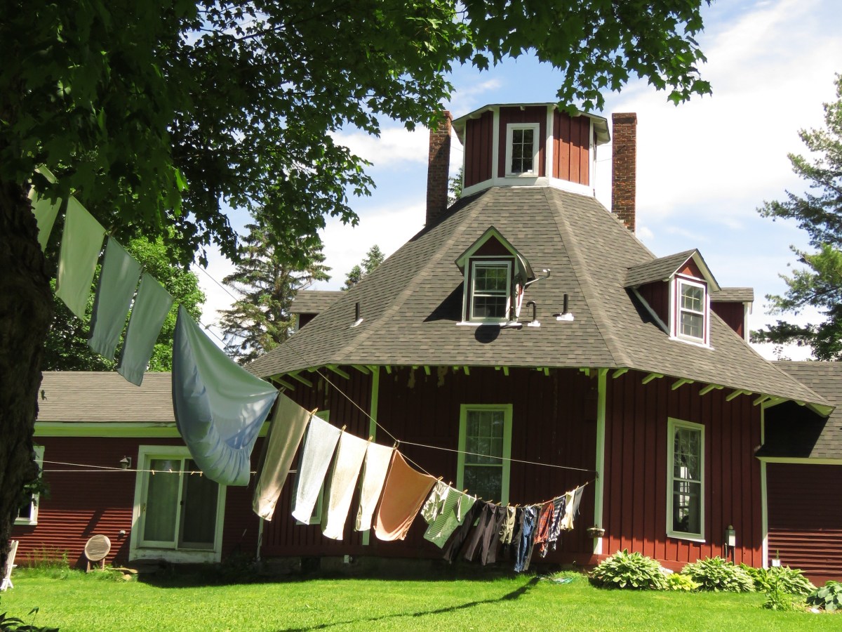 Red octagon-shaped house with tower, surrounded by green grass and trees, with clothes hanging on a line to dry in the foreground.