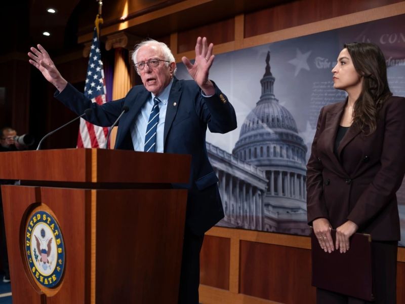 An older man speaks at a podium with raised hands while a younger woman stands beside him holding a folder; the U.S. Capitol is depicted in the background.