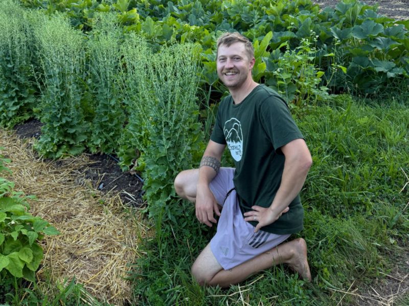 A man kneels on the grass beside tall leafy plants in a garden or farm, smiling at the camera.