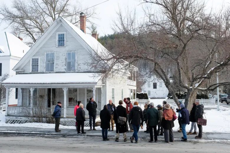 A group of people stand and talk on a snowy street in front of an old, white, wooden house with leafless trees and a picket fence.