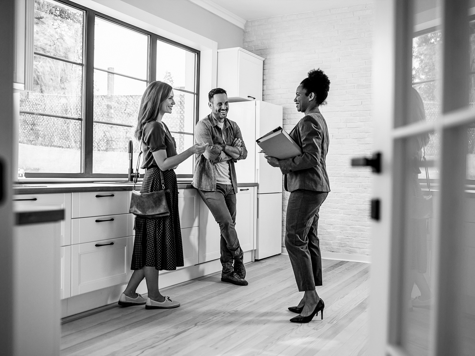 Three people stand and talk in a modern kitchen; one holds a folder, while the other two appear to listen and smile.