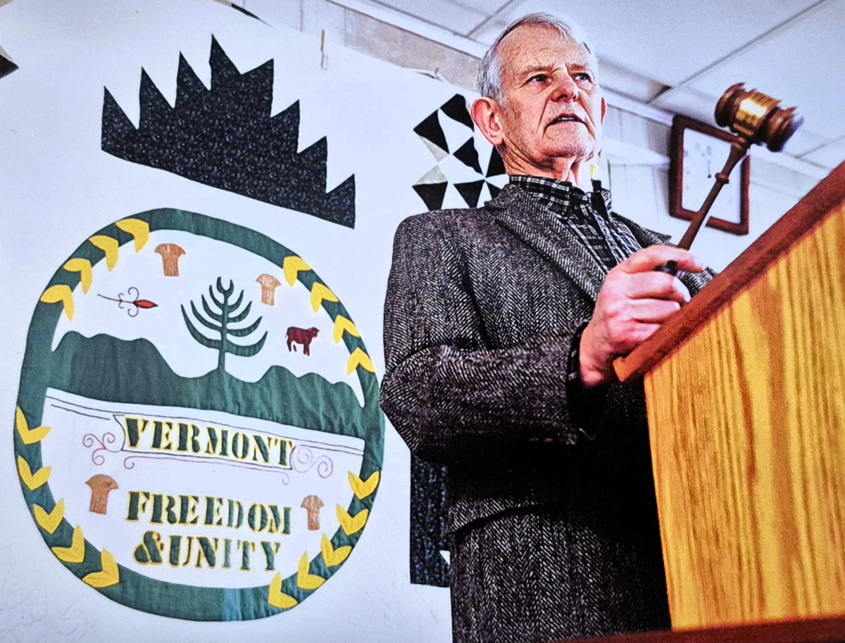 An elderly man stands at a podium holding a gavel, with the Vermont state seal and the words "Vermont Freedom & Unity" displayed on a banner behind him.